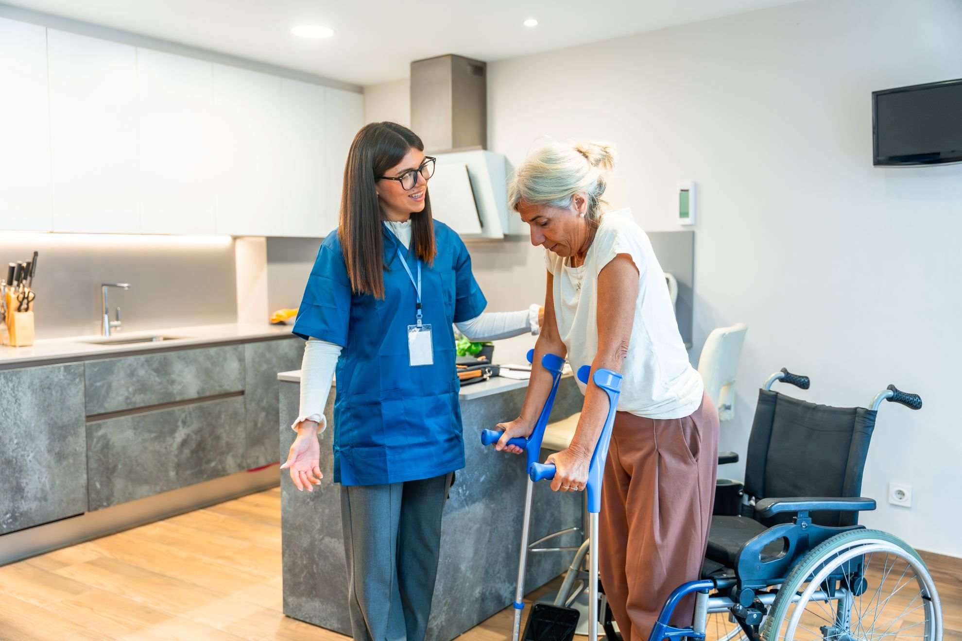 Nurse in blue scrubs supporting a senior woman walking with crutches during rehabilitation